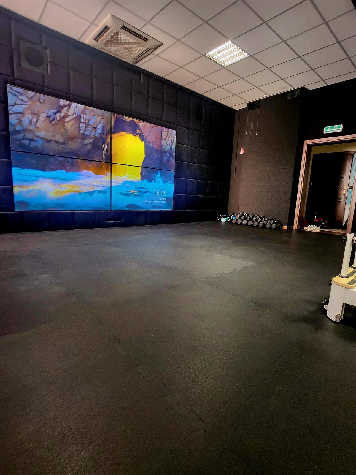 Otwarta przestrzeń treningowa z miękką wykładziną, ścianą wideo i rzędem hantli oraz kettlebelli pod ścianą