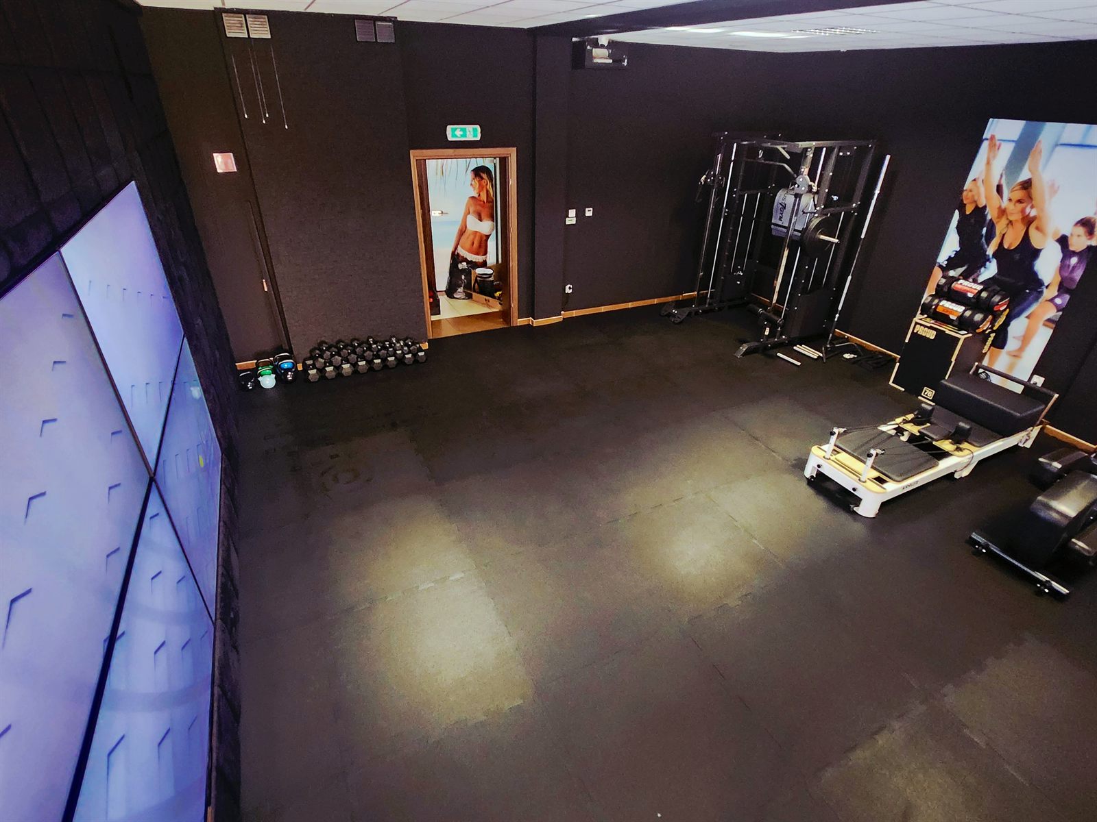 Sala treningowa BodyTop widziana z góry — po lewej ściana z ekranów, pod ścianą rząd hantli i kettlebelli, w głębi brama wielofunkcyjna i reformer do pilatesu