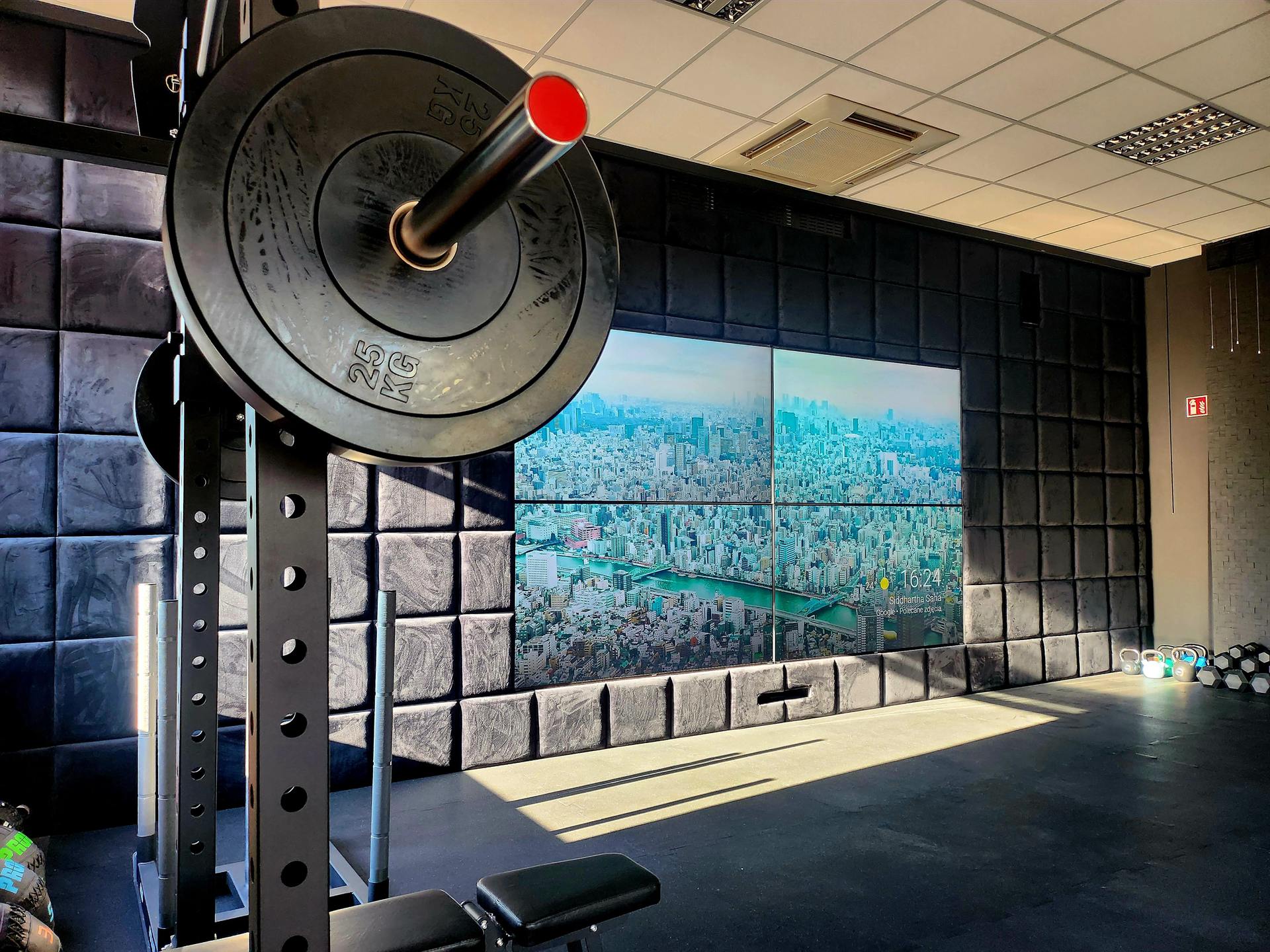 Wnętrze BodyTop Studio — sztanga z talerzem 25 kg w stojaku, w tle ściana wideo z panoramą miasta i tapicerowana ściana akustyczna